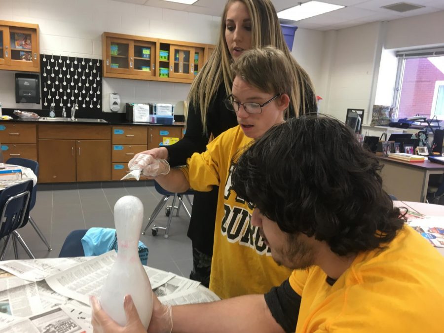 Lauren McIntyre assists students Samuel Littleton (left) and Nicolas Mejas (right) painting bowling pins for their new product, soon to be sold in the mail room and coffee shop to teachers.