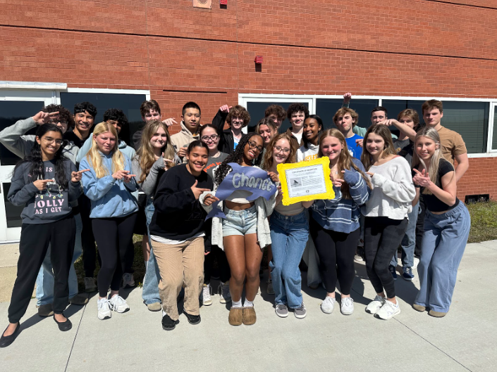 The Current veteran staff poses with a Chance the dolphin cutout and the Certificate of Adoption for the dolphin. Photo taken on March 12, 2025 outside of Ocean Lakes High School.