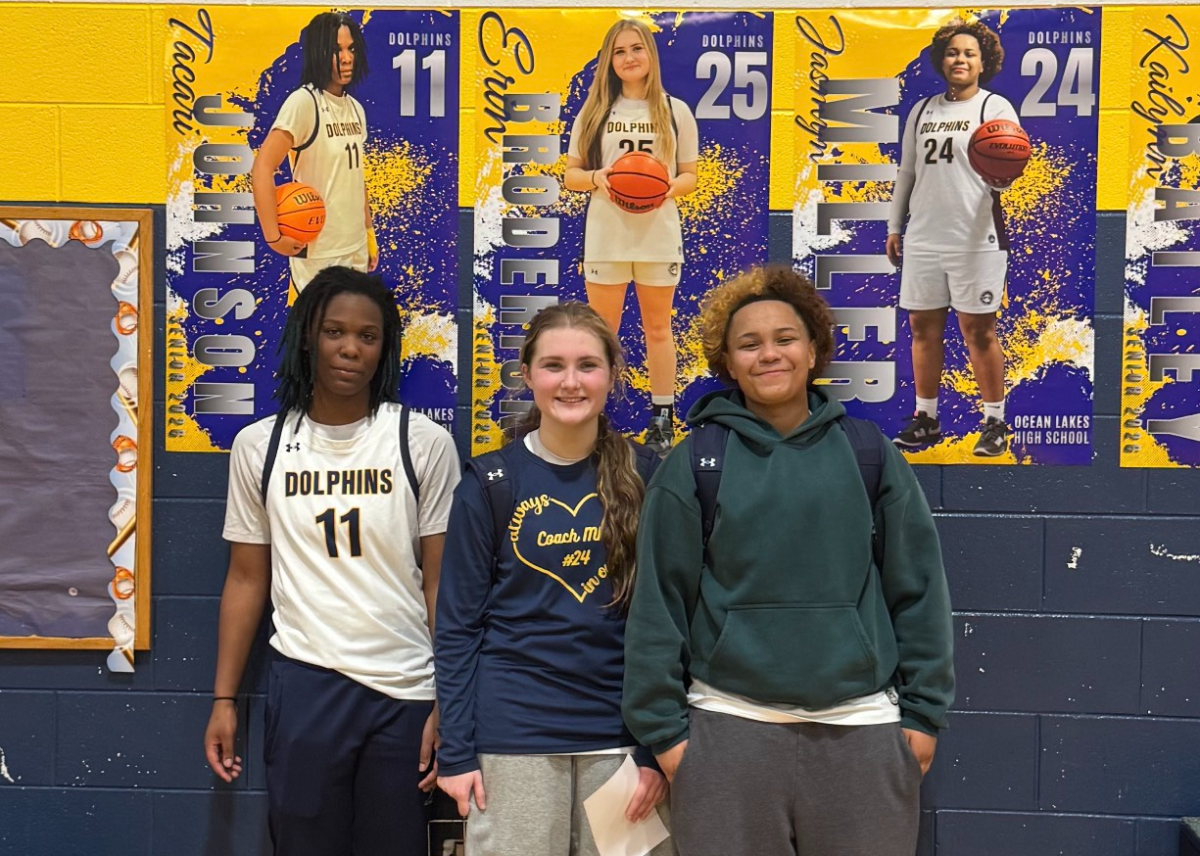 Pictured left to right: Seniors Tacari Johnson, Erin Broderick and Jasmyn Miller pose with smiles below their senior posters on senior night. Senior night occurred on Feb. 10, 2026 at Ocean Lakes High School. Photo taken by Natasha Wolmarans.
